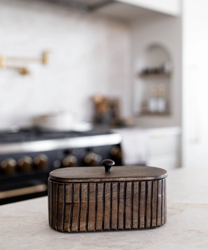 A dark wooden oval box with vertical grooves and a small round handle on top styled on a kitchen counter. The wood's texture feels rustic, conveying a warm, vintage aesthetic. | SW Home
