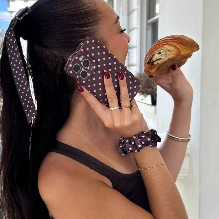 Woman with a brown polka dot scrunchie holds a pastry and talks on the phone.