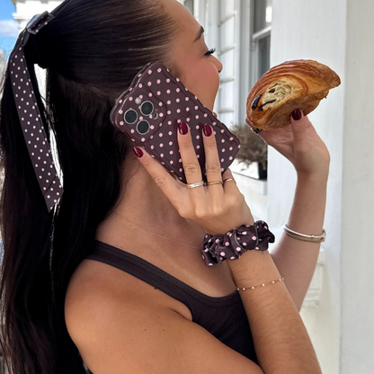 Woman with a brown polka dot scrunchie holds a pastry and talks on the phone.