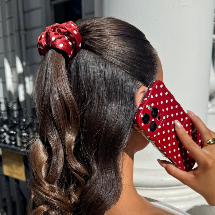 Woman with shiny brown hair wearing a burgundy polka dot scrunchie and holding a matching phone case.