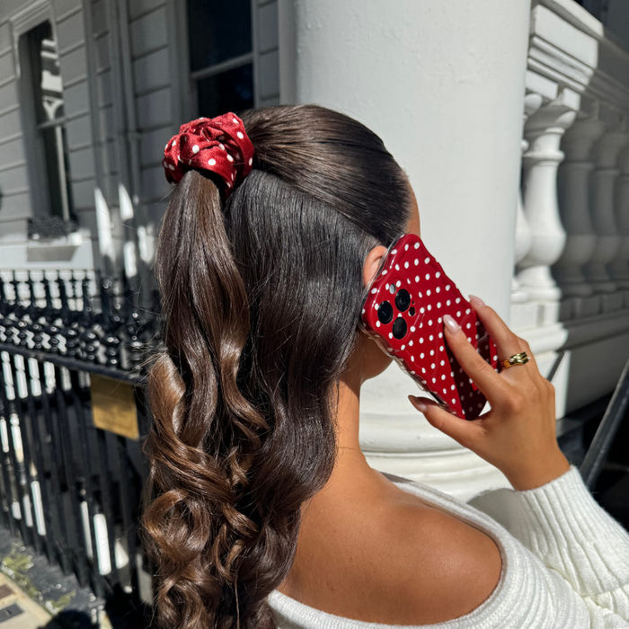 Woman with wavy brown hair wearing a burgundy polka dot scrunchie stands outside a white building.