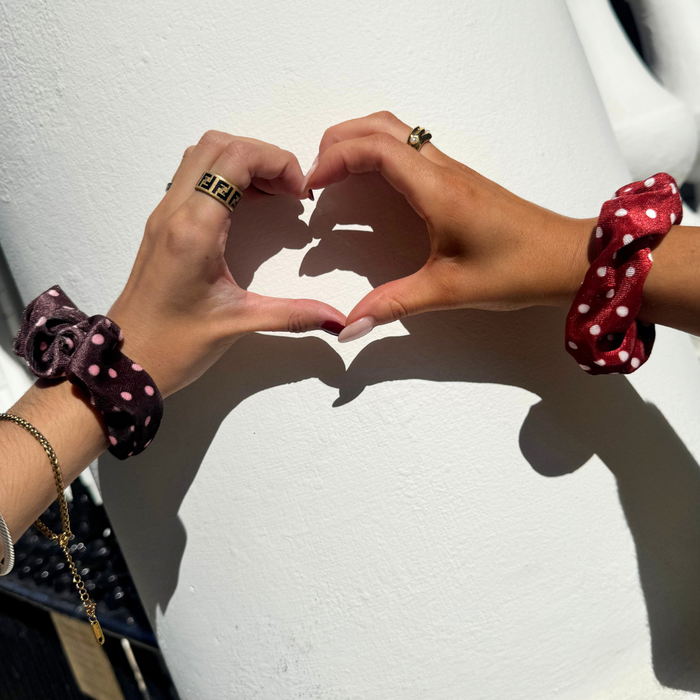 Two hands form a heart shape, each wearing a burgundy scrunchie with white polka dots in natural sunlight.