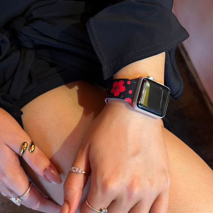Hand with rings wears a Coconut Lane Apple Watch strap in a cherry and hibiscus pattern, resting on their lap.