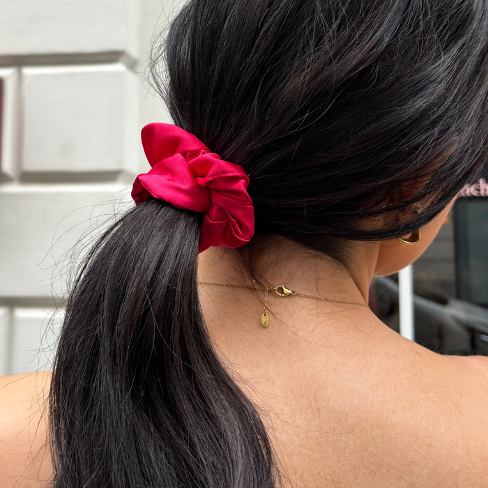Woman with dark hair in a ponytail, wearing a bright red scrunchie and gold necklace, seen from behind.