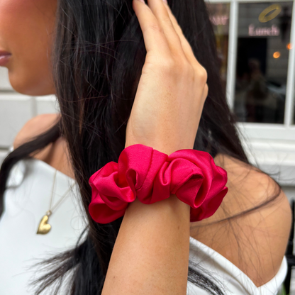 Woman outdoors wearing a vibrant red scrunchie on her wrist, white outfit, and a gold heart necklace.
