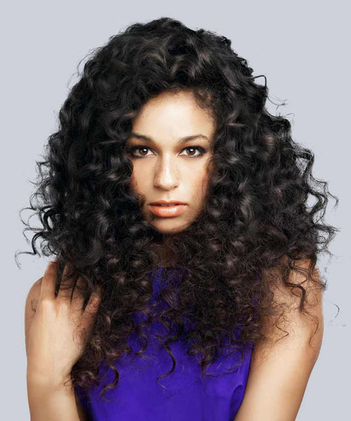 A person with long, thick, curly Natural Black  hair in a sleeveless purple top looks at the camera against a gray background.