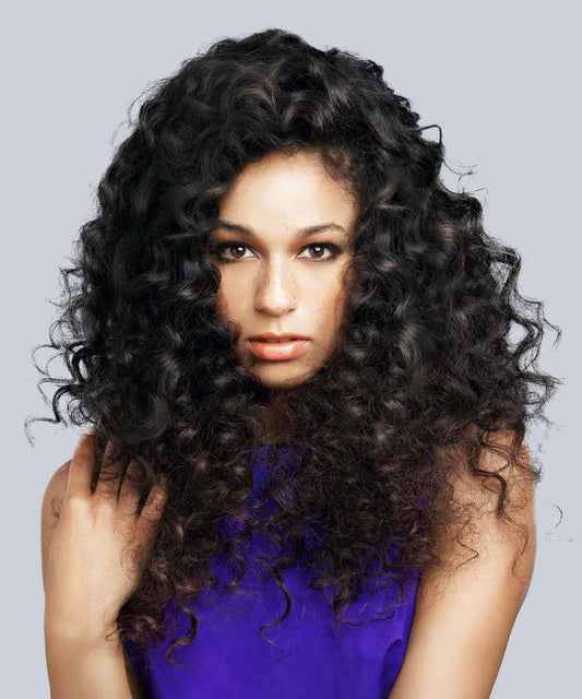 A person with long, thick, curly Natural Black  hair in a sleeveless purple top looks at the camera against a gray background.