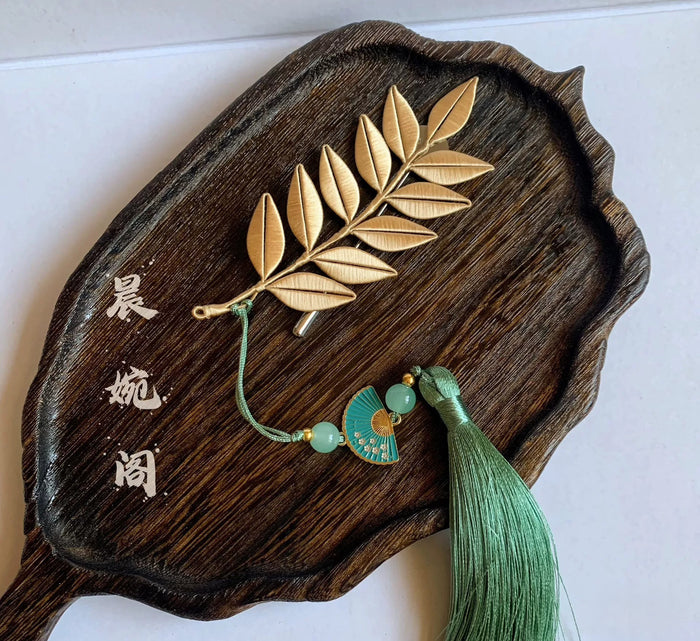 Handmade gold leaf branch brooch with green tassel and fan pendant, displayed on dark wood tray
