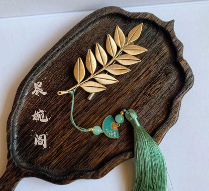 Handmade gold leaf branch brooch with green tassel and fan pendant, displayed on dark wood tray