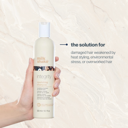 Hand holding a bottle of milk_shake Integrity shampoo against a beige background