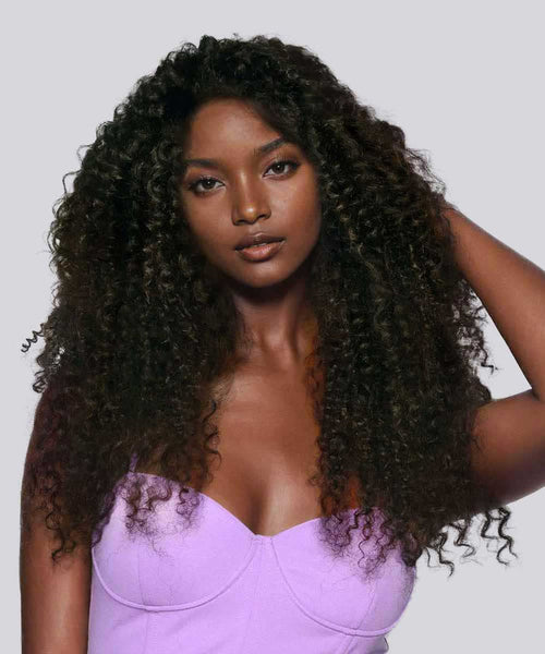 A woman with long, voluminous Perfect Locks Kinky Curly hair in Natural Black  poses against a light gray background in a strapless lavender top.