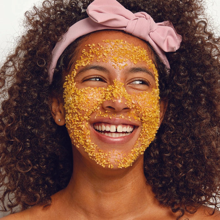 Woman smiling with golden organic face scrub, curly hair, pink headband — natural skincare routine
