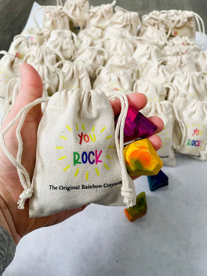 Adult hand holding  the mini beige drawstring bag that has the test YOU ROCK in rainbow letters . The Original Rainbow Crayon logo is much smaller underneath in black text. Hand is also showing a yellow and pink mini crayon rock for scale. Bag fits perfectly in the hand of an adult .