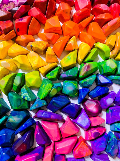 A ROY G BIV rainbow order display of multicolored crayon rocks from Art 2 the Extreme. Each crayon is in a multi-colored color family and will color in various colors. Top left of the image starts with the warmer hues such a pink, red, and orange. The middle has crayon rocks in the colors of yellow, green, then blue. The bottom is blue crayons, pink crayons, and purple crayon rocks.