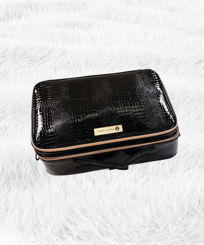 A sleek black, crocodile-patterned rectangular travel case with a gold zipper and label sits elegantly on a white, fluffy surface.