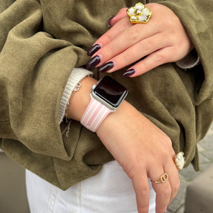 Coconut Lane Powder Stripes Apple Watch strap with pastel stripes on wrist, paired with rings and an olive jacket.
