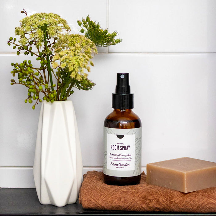 Room spray bottle with a label, soap bar, and vase with flowers on a wooden surface.