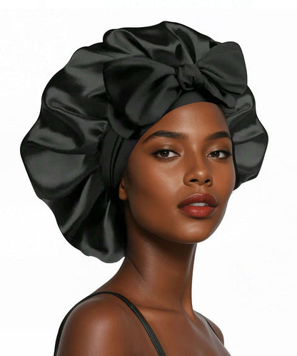 A woman with smooth brown skin wears a luxurious black satin bonnet with a big bow, looking confident against a plain white background.