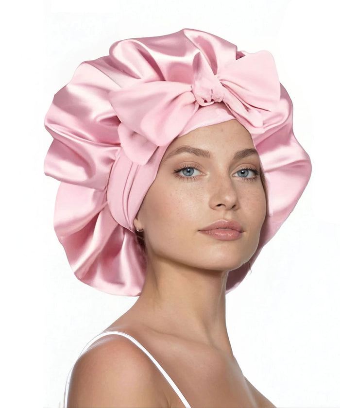 A woman wears a large, shiny pink satin bonnet with a big bow on the front, confidently gazing at the camera against a white background.