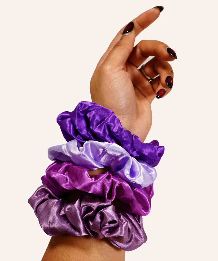 A hand with black and red nails is raised, wearing four satin scrunchies in purple, lavender, and light purple on the wrist.