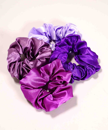 Four satin scrunchies in lavender, lilac, deep violet, and magenta cluster on a light surface, adding style to any collection.