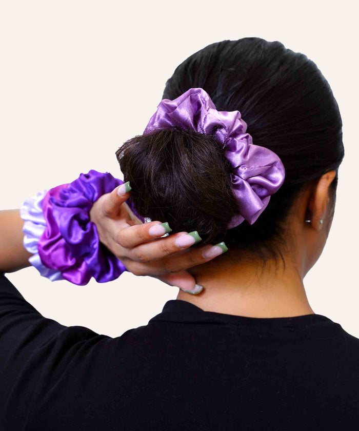 A person with dark hair in a bun wears a shiny purple scrunchie, holding their head, with two scrunchies on their wrist. Stylish background.