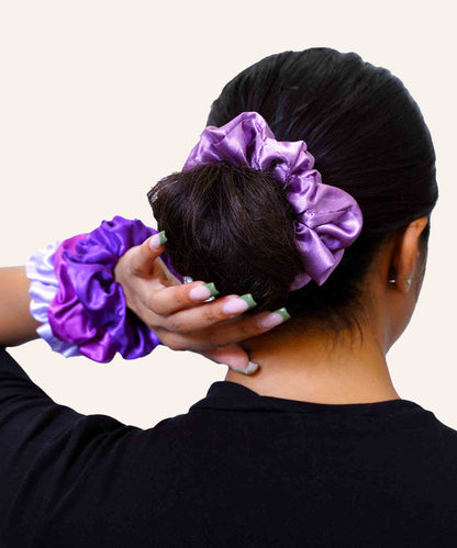 A person with dark hair in a bun wears a shiny purple scrunchie, holding their head, with two scrunchies on their wrist. Stylish background.