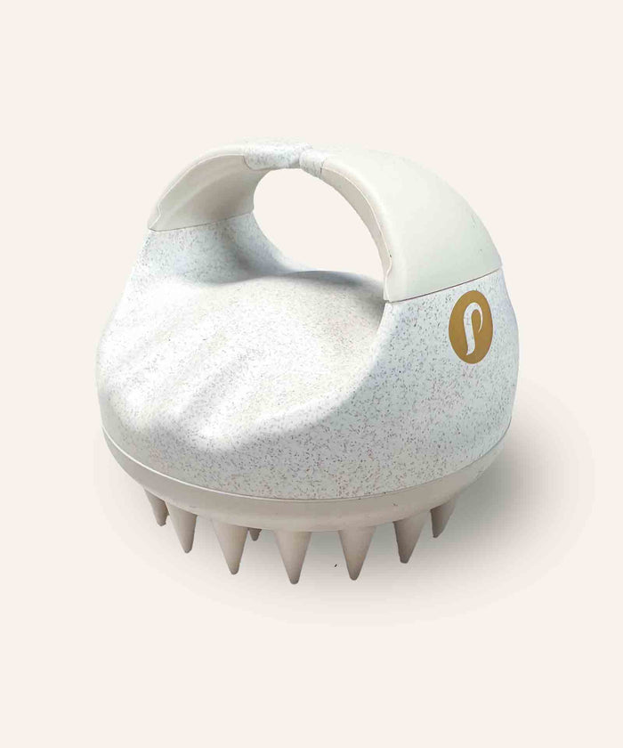 A handheld scalp massager with a curved handle, white speckled body, beige rubber bristles, and a gold logo for scalp care.