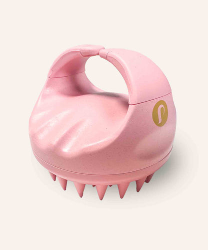 A pink silicone scalp massager with a handle and short, flexible bristles is designed for gentle exfoliation, on a light background.