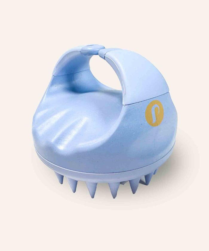 A light blue handheld scalp massager with soft bristles and a curved handle, yellow logo on the side, rests on an off-white background.