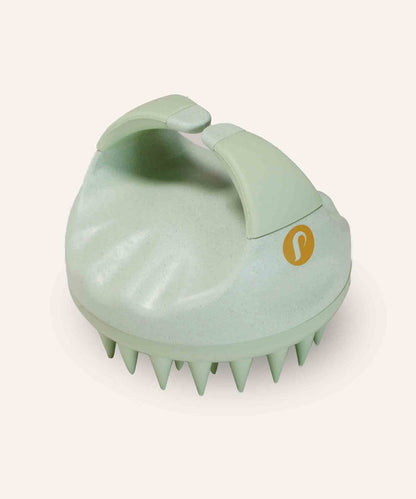 A light green silicone scalp massager with an ergonomic handle and flexible bristles for hair care, on an off-white background.