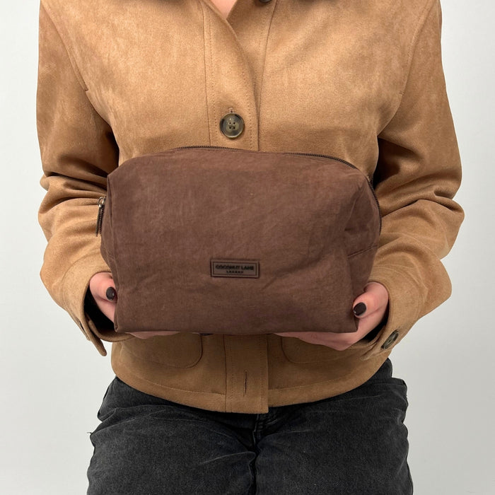 Chocolate brown suedette makeup bag held by someone in a tan suede jacket and black jeans.