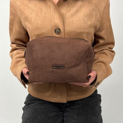 Chocolate brown suedette makeup bag held by someone in a tan suede jacket and black jeans.