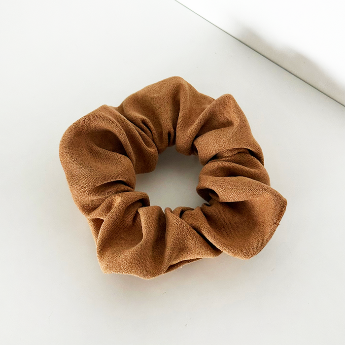Tan suedette scrunchie by Coconut Lane on a white surface near a wall corner.
