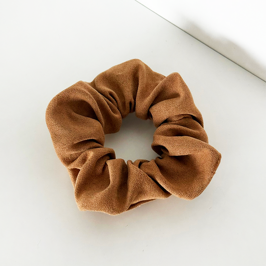 Tan suedette scrunchie by Coconut Lane on a white surface near a wall corner.