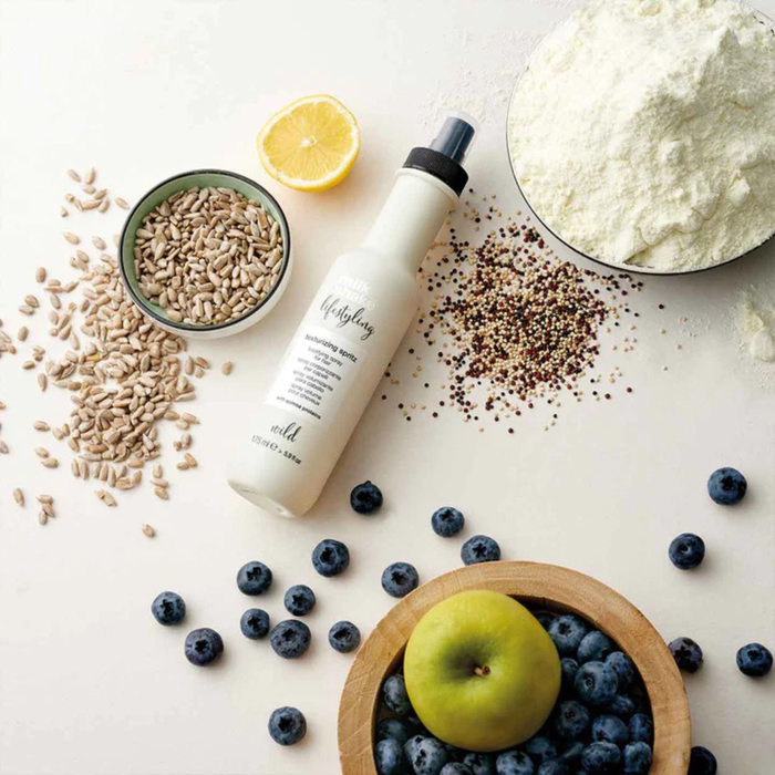 milk_shake texturizing spritz surrounded by ingredients including nuts, a lemon, and fruits on a light surface.