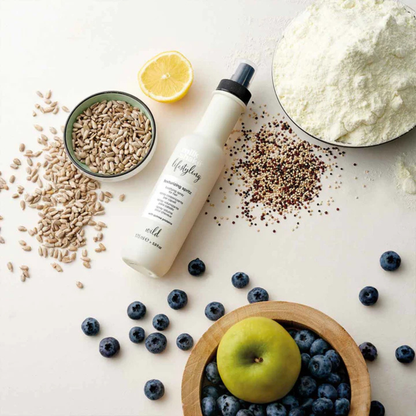 milk_shake texturizing spritz surrounded by ingredients including nuts, a lemon, and fruits on a light surface.