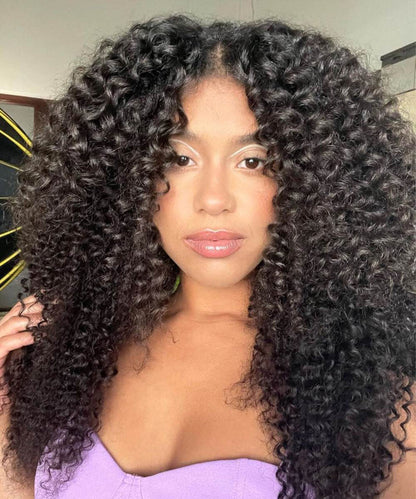 A woman with voluminous, curly dark hair poses indoors in a strapless lavender top, looking confidently at the camera.