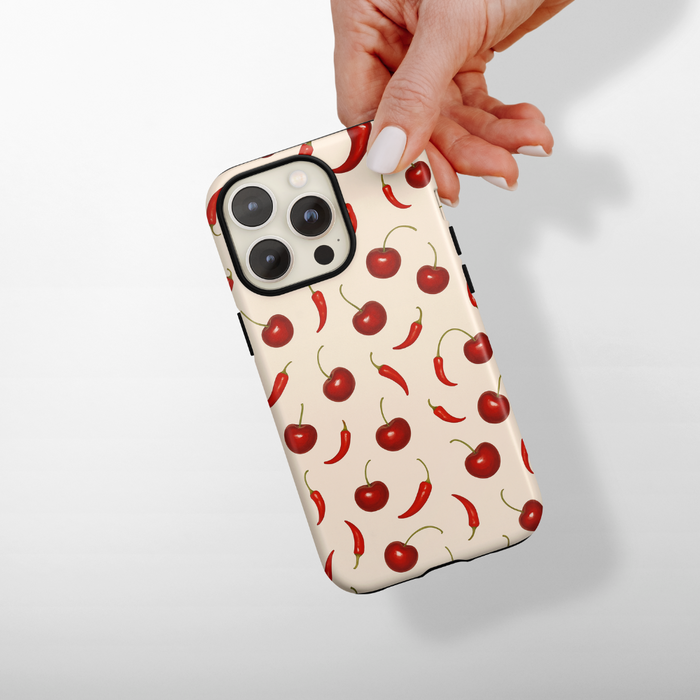 Coconut Lane Tough Phone Case in Cherry Spice design with red cherries and chili peppers on a cream background, held in a hand.
