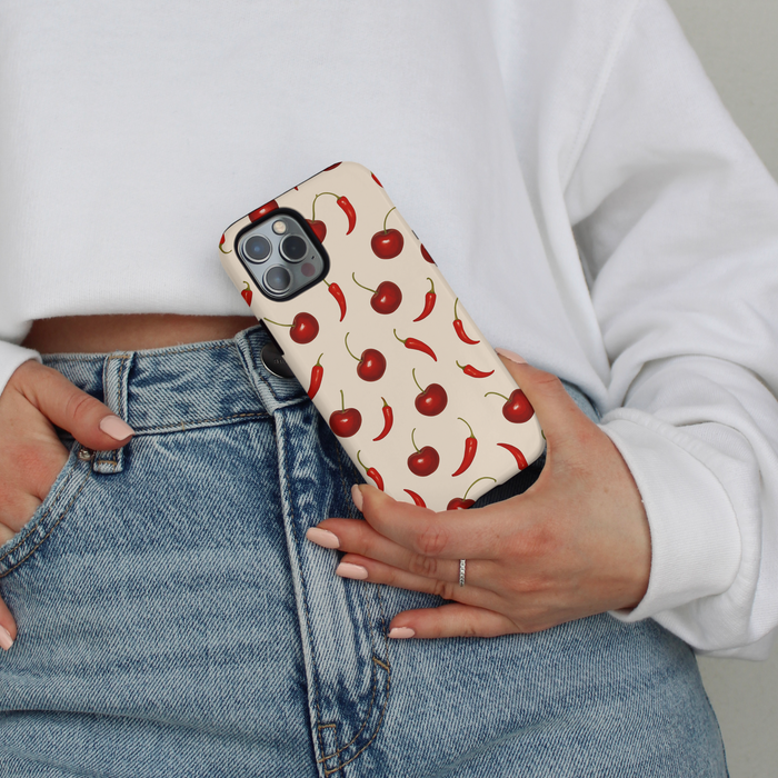 Person in jeans and a white sweater holds a Coconut Lane phone case with red cherry and chili pepper pattern.