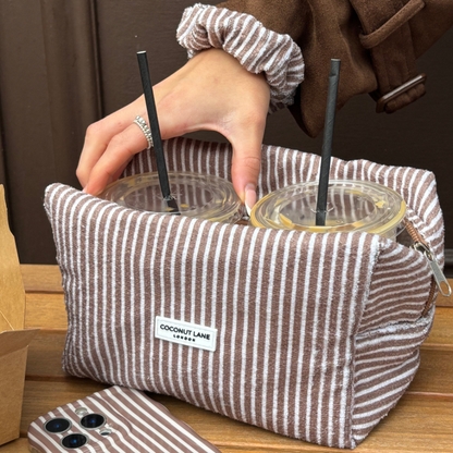 Chestnut striped Coconut Lane towel makeup bag with two iced drinks, held by a hand on a wooden table.