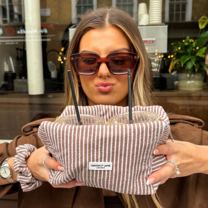 Woman in sunglasses holds two iced drinks and a Chestnut Stripes Coconut Lane towel makeup bag, smiling playfully.