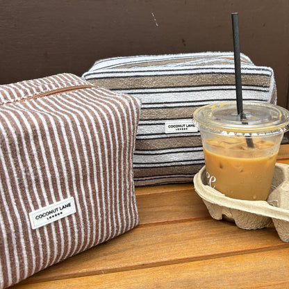 Two Coconut Lane makeup bags with retro stripe patterns beside an iced coffee on a wooden surface.