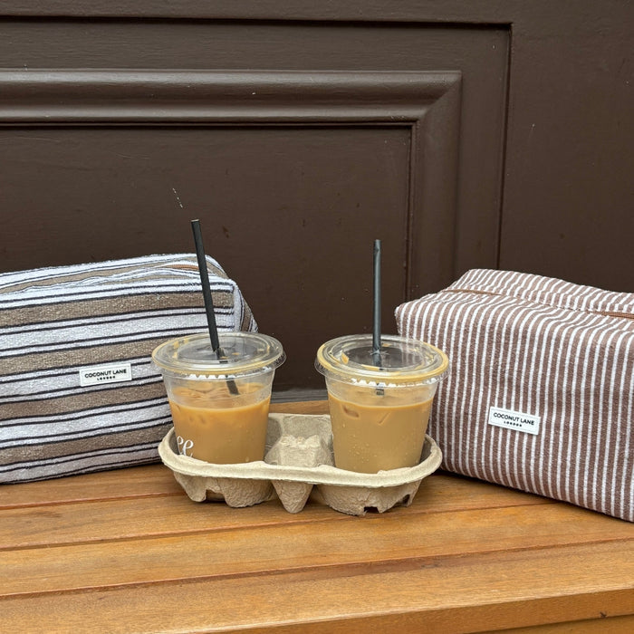 Two iced coffees on a bench between two striped Coconut Lane towel makeup bags in a cardboard holder.