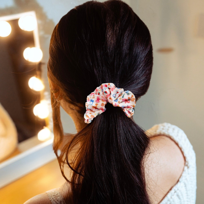 Woman with dark hair uses a white towel scrunchie to style a low ponytail, standing by her vanity.