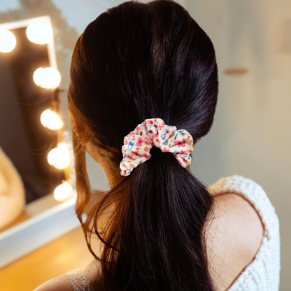 Woman with dark hair uses a white towel scrunchie to style a low ponytail, standing by her vanity.