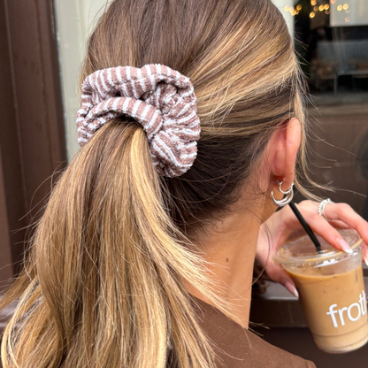 Blonde woman with Coconut Lane chestnut striped towel scrunchie, holding an iced coffee with a black straw.