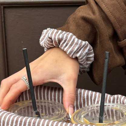 Hand with silver ring holds iced drinks in a brown and white striped Coconut Lane towel scrunchie carrier.