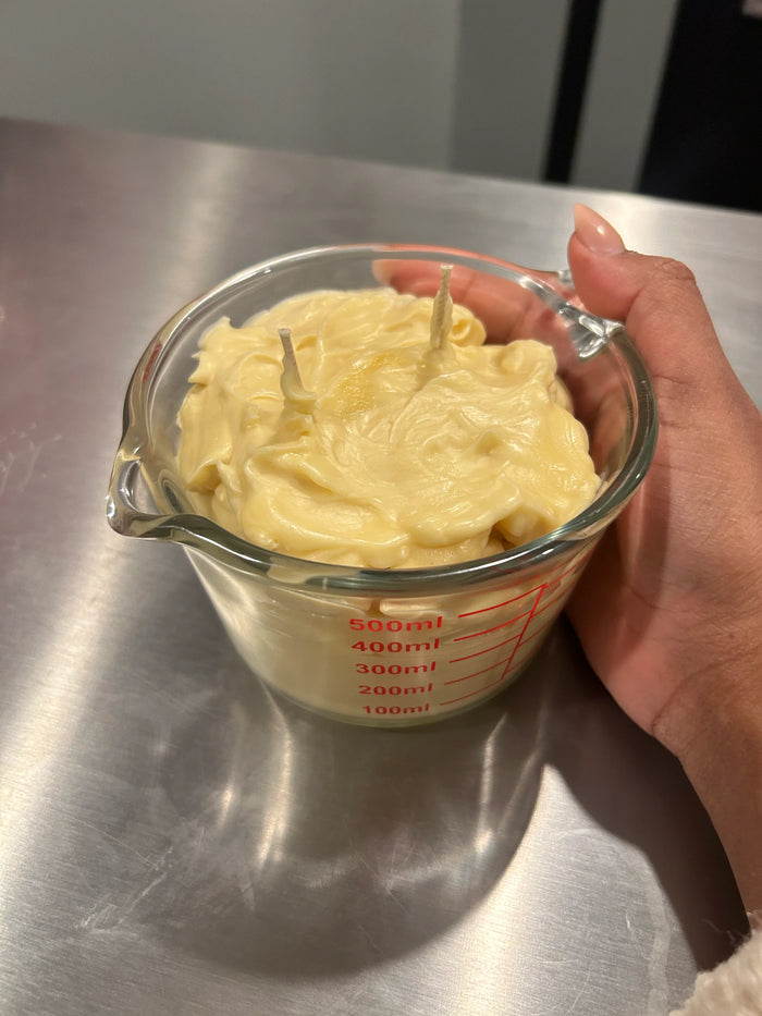 Hand holding whipped body butter in a glass measuring cup on a metal surface