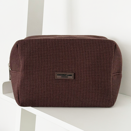Chocolate brown waffle-textured makeup bag with zip and label on a white shelf.